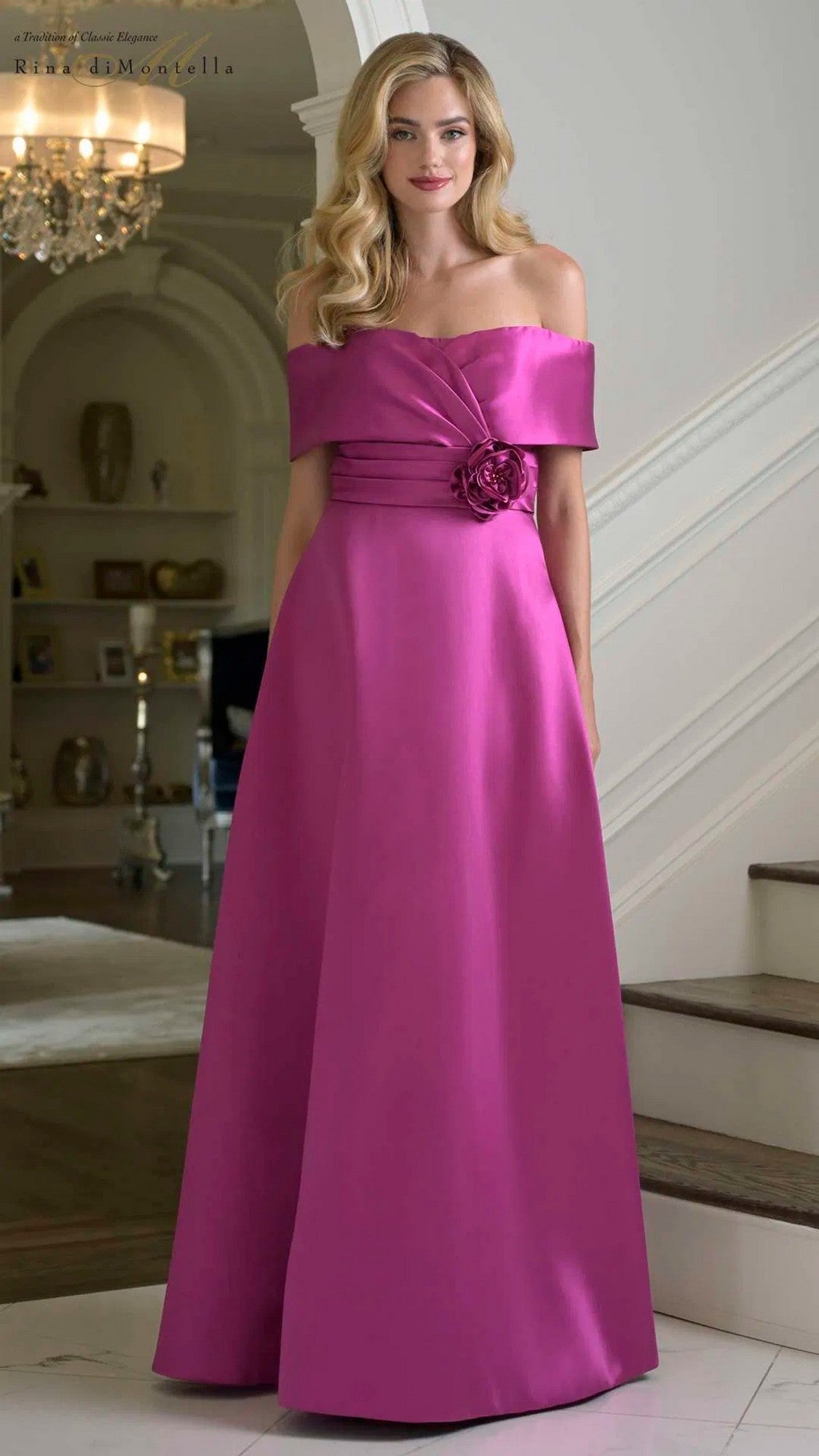 A woman stands indoors wearing the Rina Di Montella RD3429 long formal dress in magenta, an off-the-shoulder gown with floor-length Mikado fabric and a floral accent at the waist.
